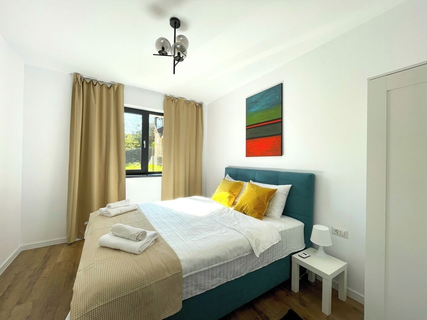 A modern bedroom with a teal headboard, a large window with curtains, a bedside table with a lamp, and a colorful abstract painting on the wall.
