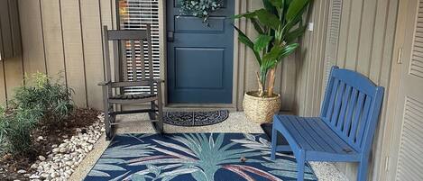 Un portico con una porta blu, una panchina, una pianta in vaso e un tappeto decorativo.