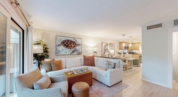 A modern living room with a white sofa, a coffee table, and a large painting of a turtle. There is a kitchen in the background with wooden cabinets and a dining area.