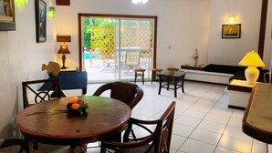 A dining area with a round table, chairs, and a fruit bowl. There is a sofa and a lamp in the background.