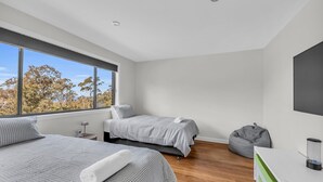 Un dormitorio moderno con dos camas, un ventanal grande, un sillón orejero gris y un televisor sobre una mesa blanca.