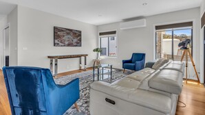 Una sala de estar moderna con un sofá grande, un sillón azul, una mesa de centro de vidrio y un cuadro en la pared.