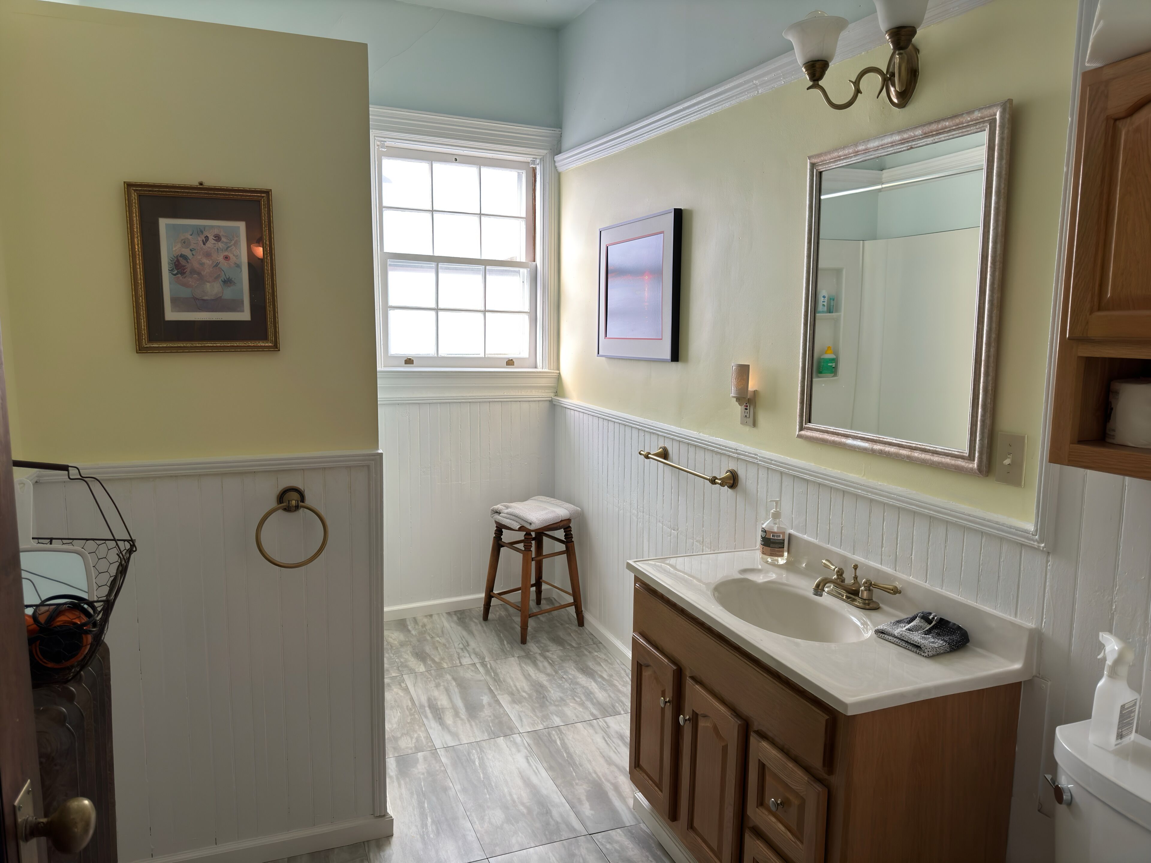 Un baño con tocador, lavabo y espejo. Hay una ventana con contraventanas blancas, un perchero y un cuadro en la pared.