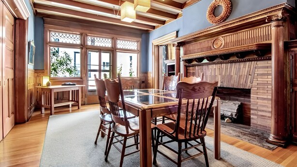 A dining area with a wooden table and chairs, a fireplace, and large windows with a view of greenery outside.