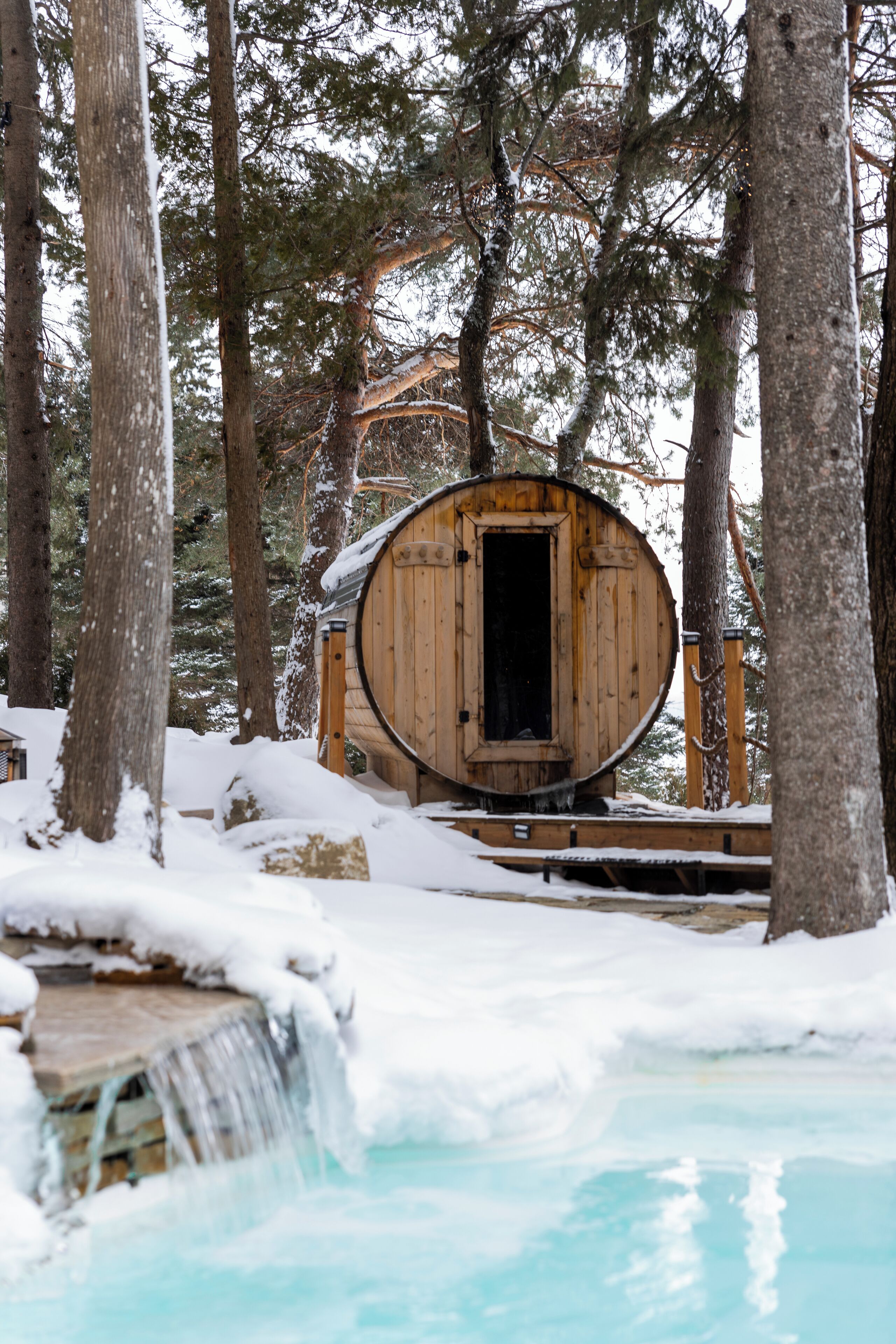 Una caba ña de madera con una puerta, rodeada de árboles cubiertos de nieve y una cascada pequeña.