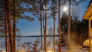 A serene outdoor setting with a hot tub surrounded by stone pathways and snow-covered ground, overlooking a lake and forested area. - Estello Suites et Spa (Frontenac)