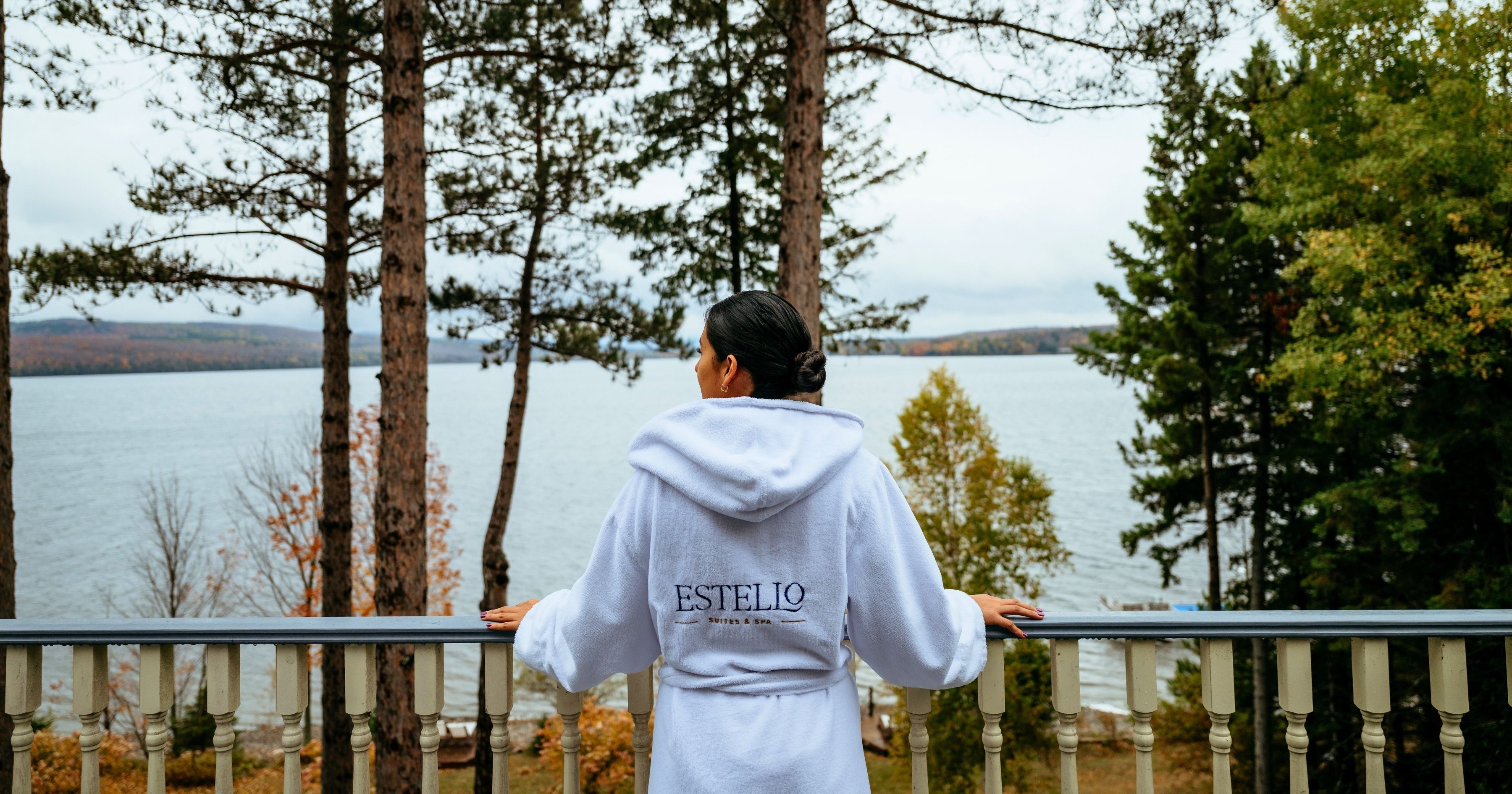 Eine Person steht auf einem Balkon mit Blick auf einen See, Bäume und ferne Hügel.