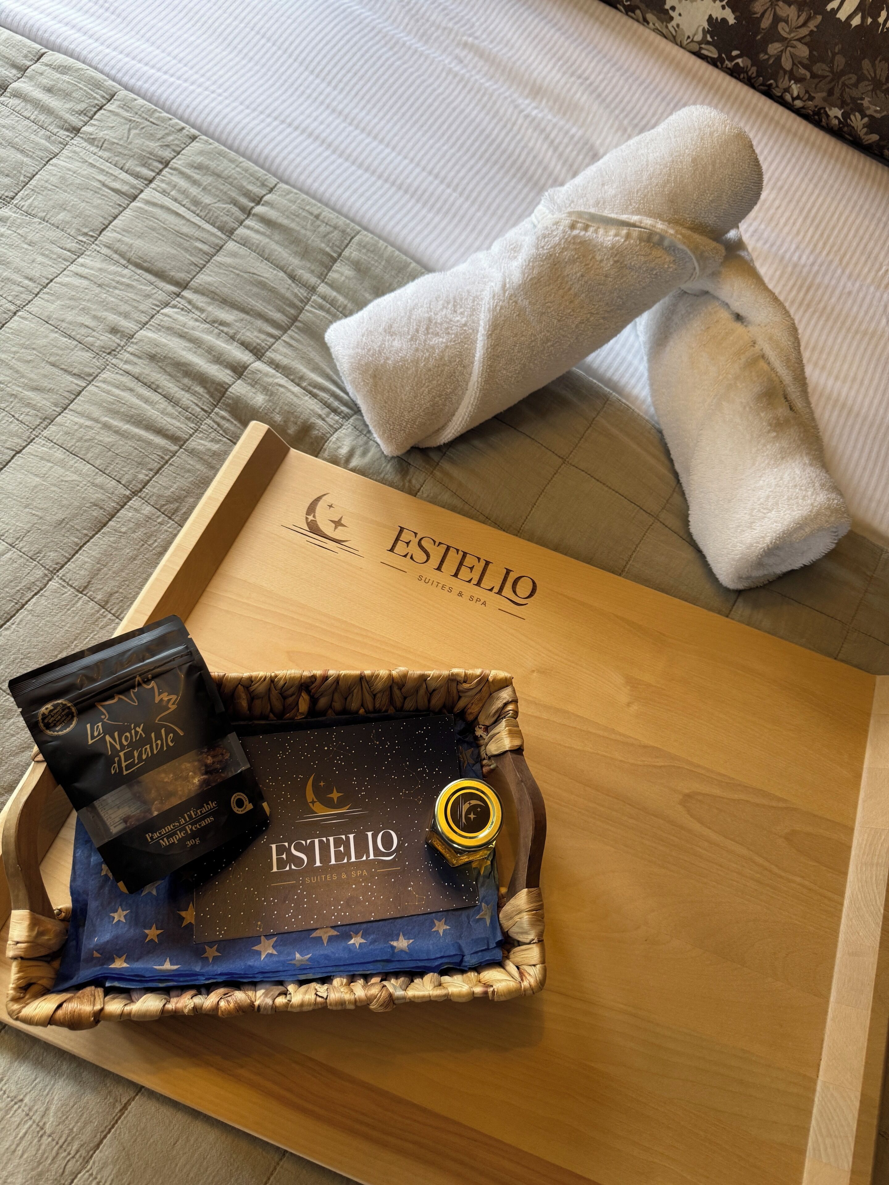 Una bandeja de madera con un paño azul estampado con un motivo de noche estrellada, una botella de Estelio Suites & Spa y un paquete de La Noix d'Erable sobre una cama con cubrecama acolchado y una toalla blanca.