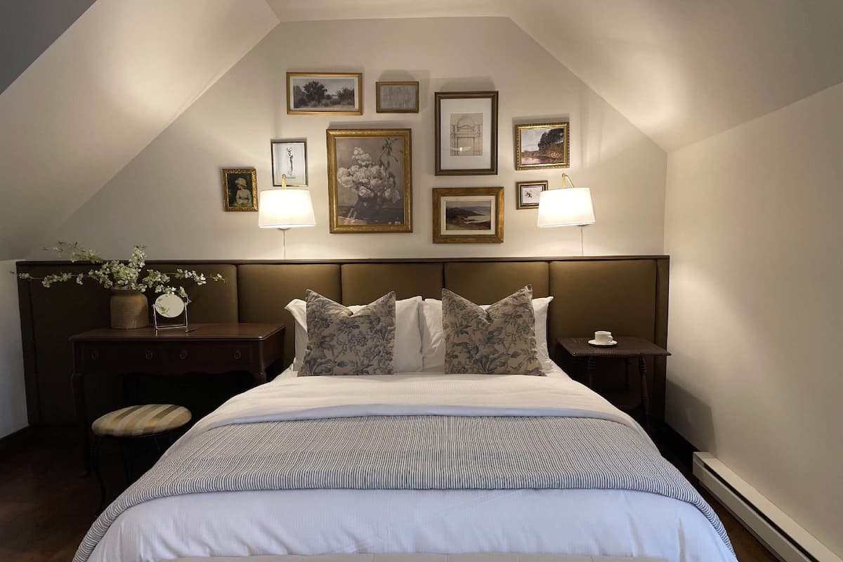 A neatly arranged bedroom with a large bed, two bedside tables with lamps, a small table with a vase of flowers, and a bench.