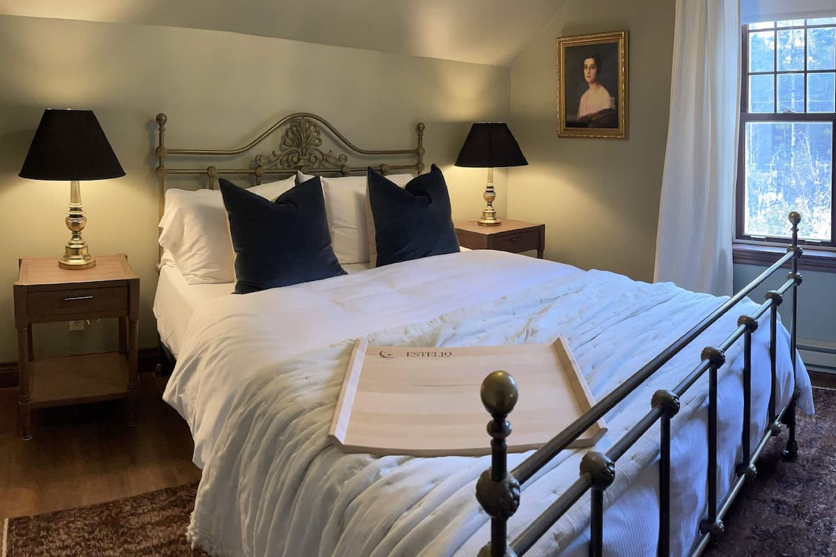 A neatly made bed with a metal headboard, two bedside tables with lamps, a framed portrait on the wall, and a window with curtains.