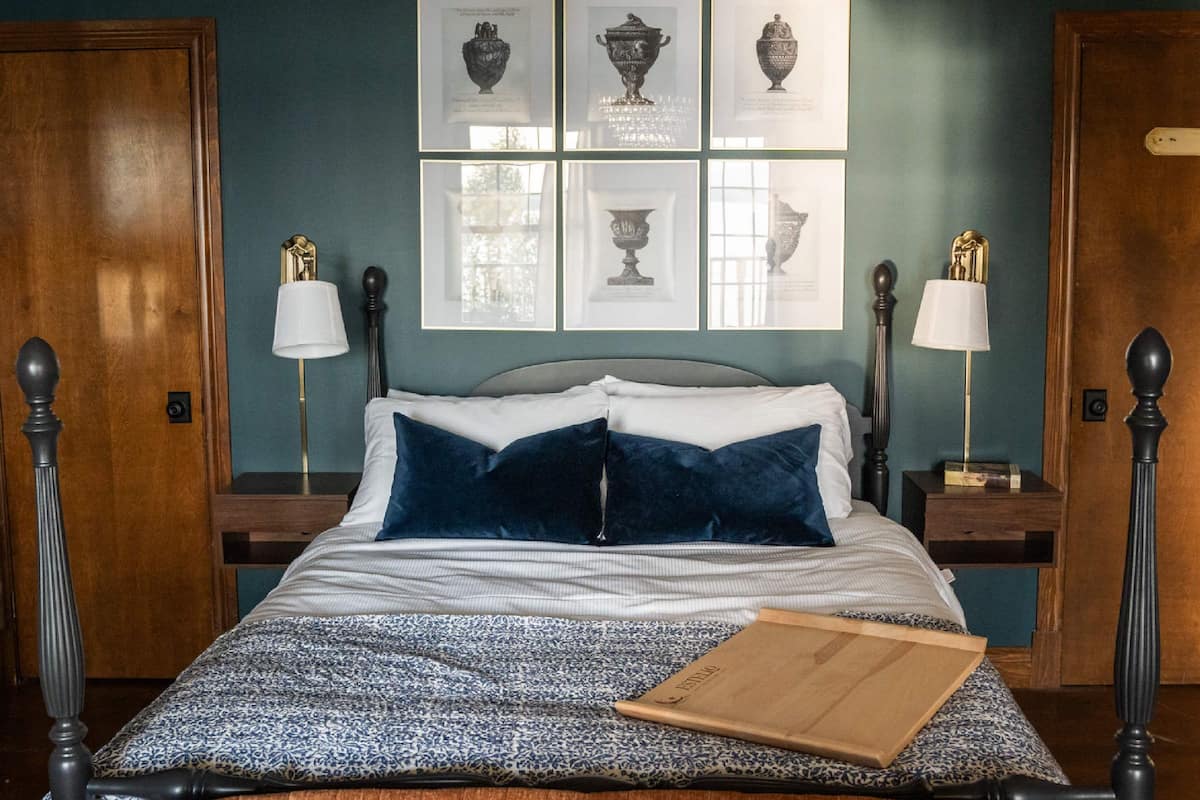 A neatly made bed with a patterned bedspread, two pillows, and a wooden tray on the bed. There are framed pictures on the wall above the bed, and two bedside tables with lamps.