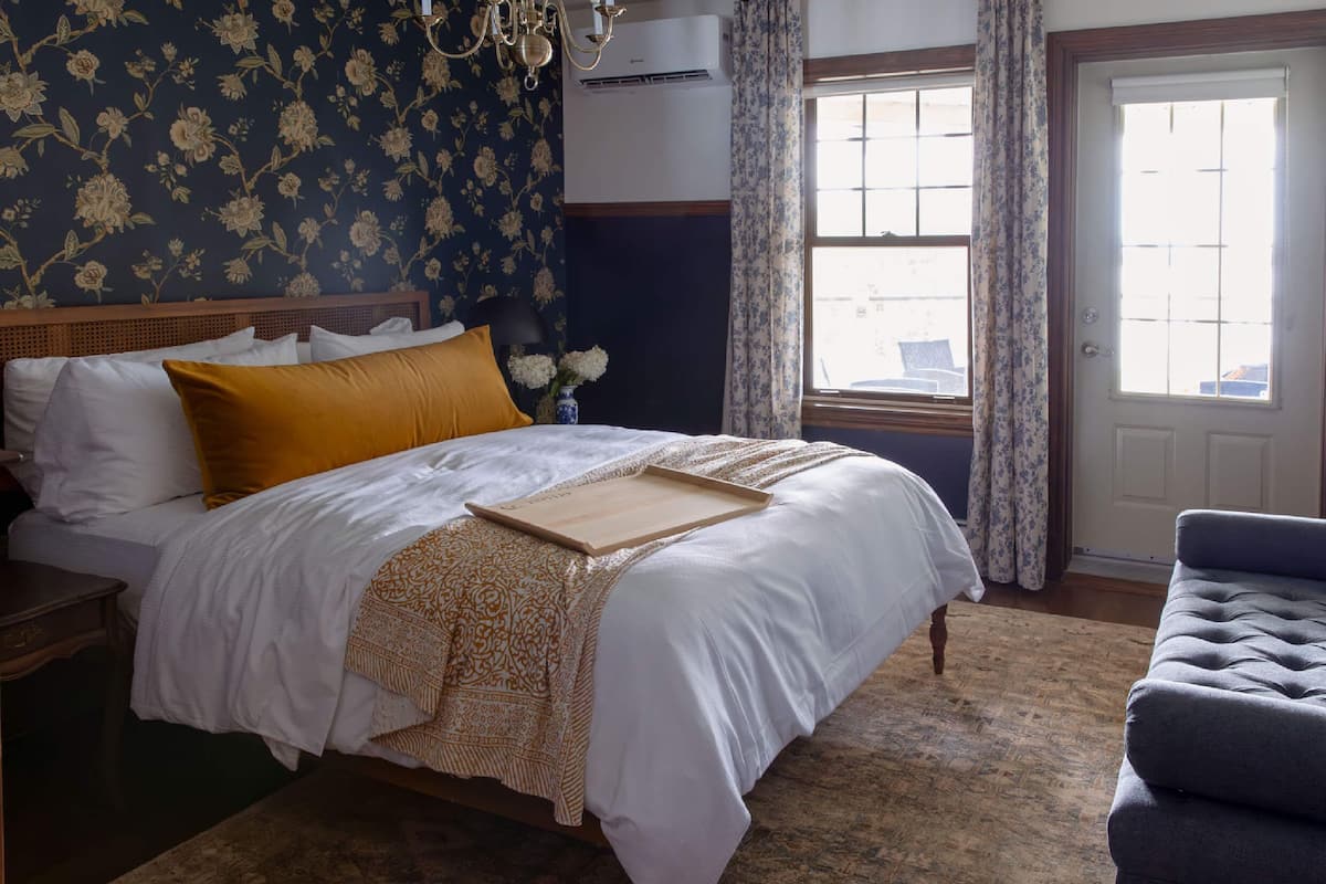 A bedroom with a large bed, a wooden nightstand, a chair, a window with curtains, and a floral wallpaper.