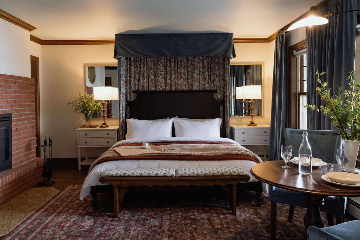 A hotel room with a large bed, a bedside table with lamps, a small table with glasses and plates, a fireplace, and a window with curtains.