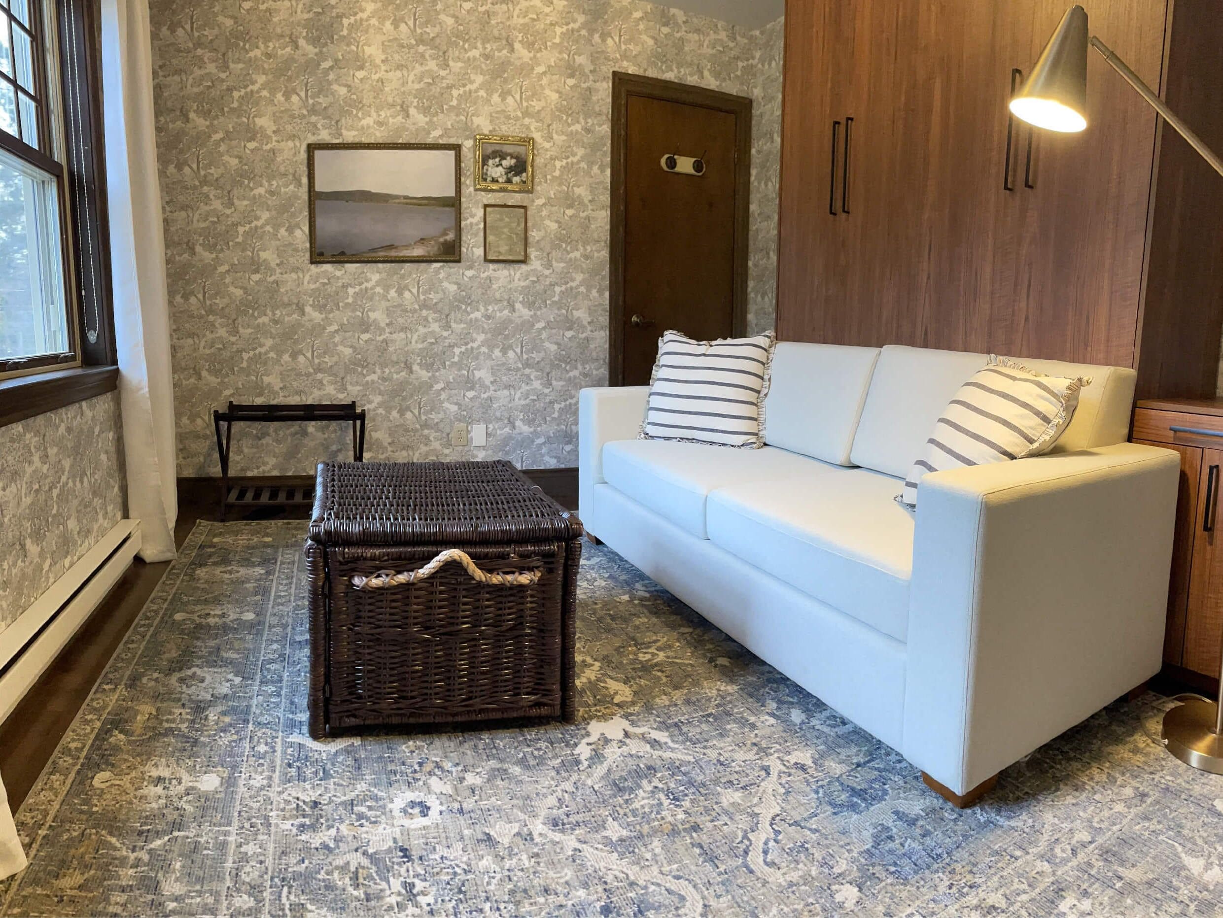 Quarto com sofá branco, um baú de vime, tapete estampado e porta de madeira.