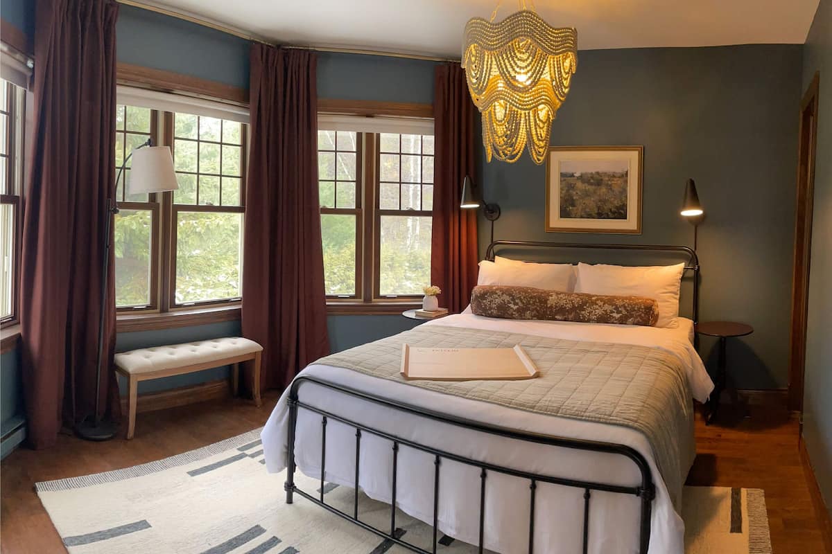 A bedroom with a bed, bench, bedside table, lamp, and a decorative hanging light fixture.