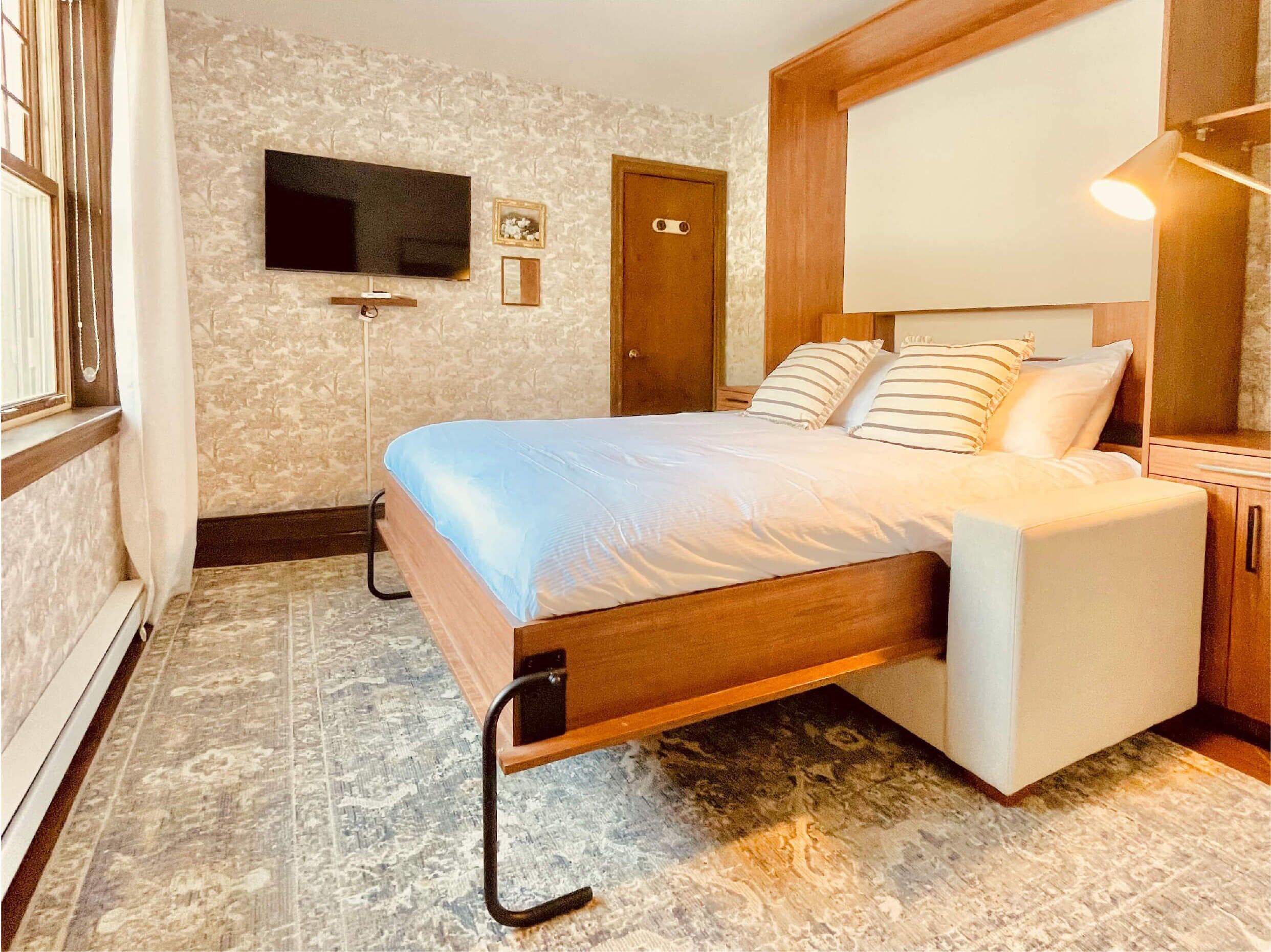 A neatly arranged bedroom with a bed, a television mounted on the wall, a door, and a bedside table.