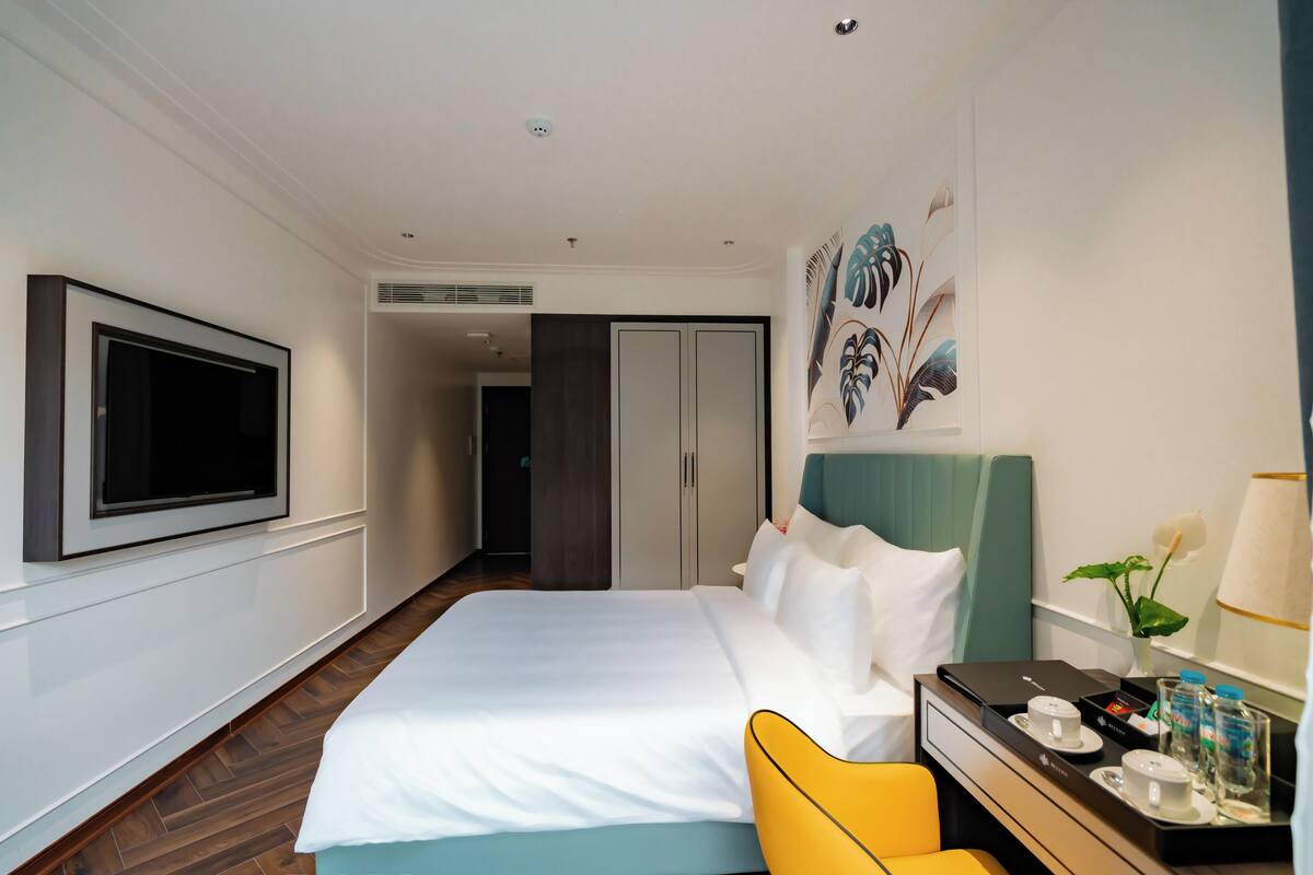 A hotel room with a bed, a TV mounted on the wall, a desk with a tray of amenities, and a door leading to another room.