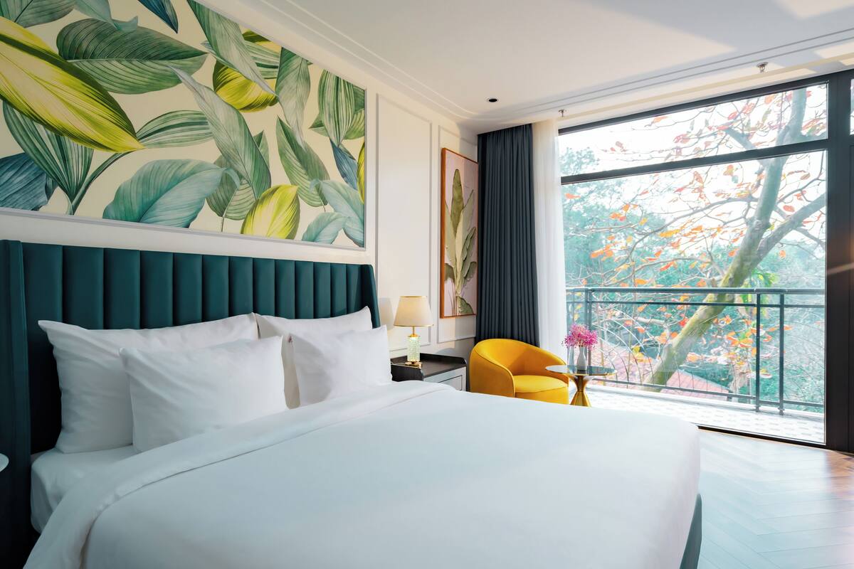 A hotel room with a large bed, a view of trees through the window, and a tropical leaf mural on the wall.