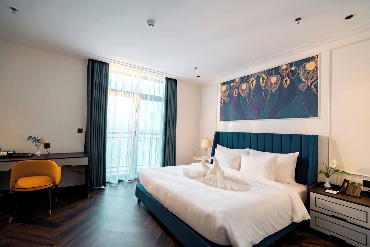 A hotel room with a large bed, a desk, a chair, and a painting on the wall.