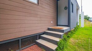 Een modern gebouw met horizontale beplating, een trap van beton die naar een deur leidt, en een kleine tuin met gras en struiken.
