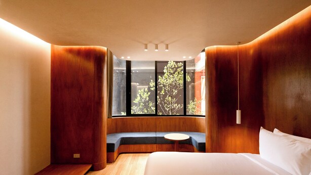 A modern hotel room with a large bed, a built-in bench with a round table, and a large window offering a view of trees. - Monder House (Chiang Mai)