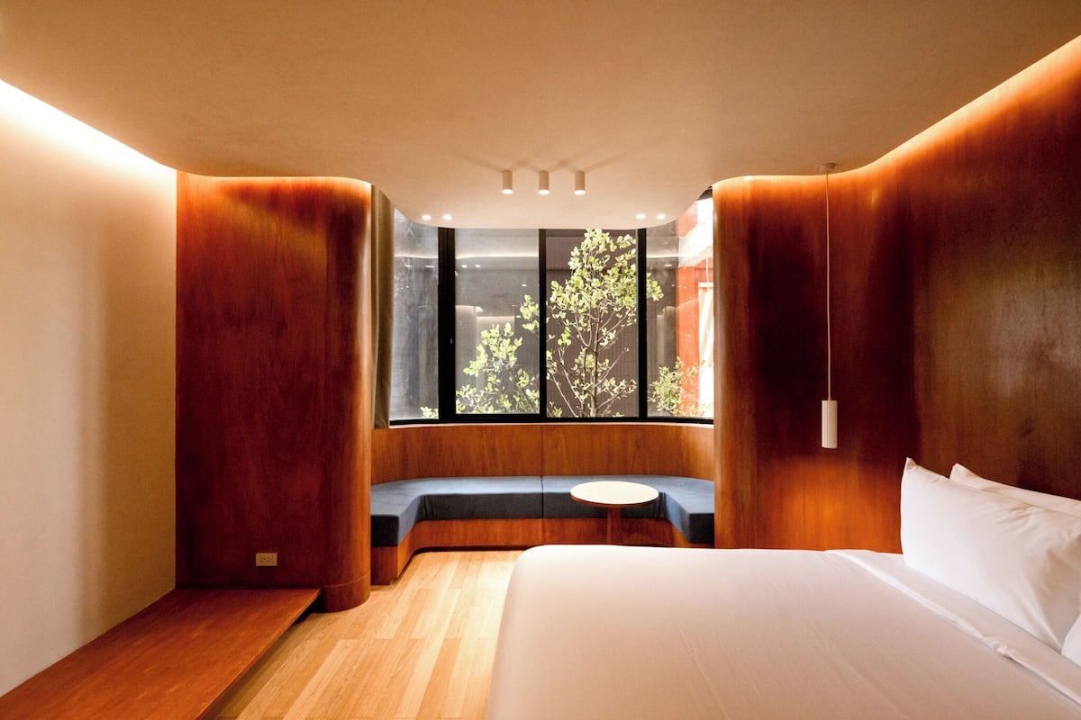 A modern hotel room with a large bed, a built-in bench with a round table, and a large window offering a view of trees.