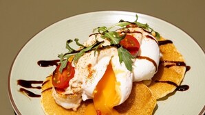 A plate with a poached egg, tomato, and greens on a bed of pancakes, drizzled with sauce. - Monder House (Chiang Mai)