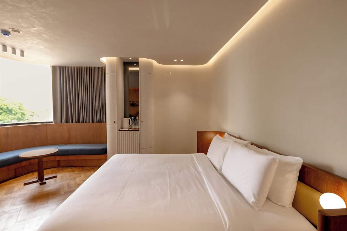 A modern hotel room with a large bed, a seating area with a table, and a view of greenery outside.