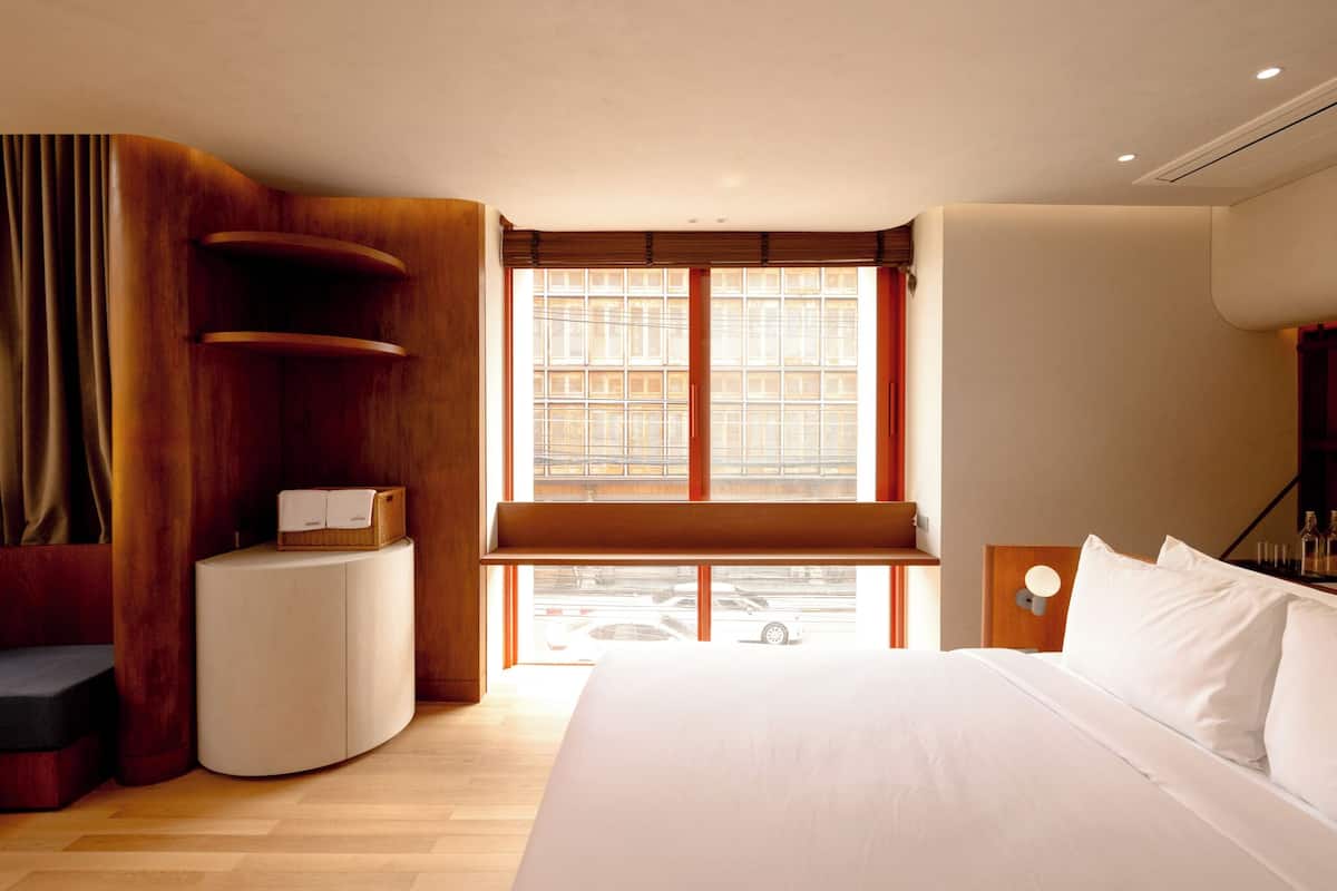 A modern hotel room with a large bed, a curved wooden sideboard, a small round table, and a large window with a view of the street.