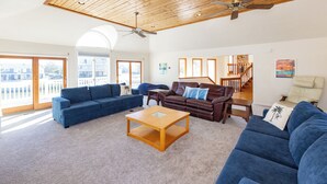 Uma sala de estar espaçosa com teto alto, um ventilador de teto de madeira, um sofá azul, um sofá de couro marrom e uma poltrona bege.