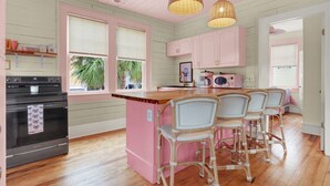 A kitchen with a pink island, wicker chairs, and a black stove. - 4 BDR Heated Pool, Professionally Designed Vacation Rental Across from Beach (Tybee Island)