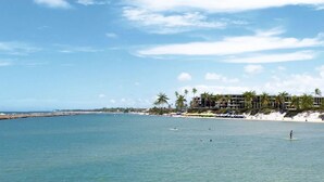 A beach with a hotel in the background, palm trees, and clear blue water. - SMART APARTMENT - EXTERNAL VIEW (Ipojuca)