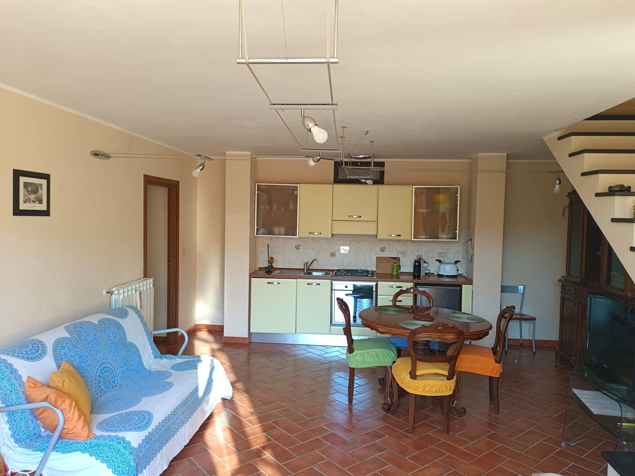 Ein Wohnzimmer mit Sofa, einem Esstisch mit Stühlen, einer Küchenzeile mit Schränken und Geräten sowie einer Treppe, die eine weitere Ebene hinaufführt.
