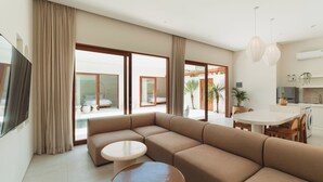 A modern living room with a beige sectional sofa, a round coffee table, and a dining area with a white table and chairs. - Villa Sari 2 by Alfred in Bali (Pecatu)