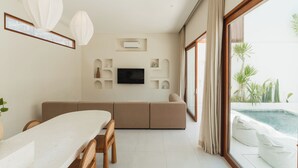 A modern living room with a large TV, beige sofa, dining table, and chairs. There is a large window with a view of a pool and plants. - Villa Sari 2 by Alfred in Bali (Pecatu)