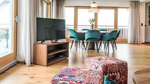 A spacious living room with a large TV, teal chairs, a wooden coffee table, and a colorful ottoman. - Panorama Dachstein by Interhome (Rohrmoos)