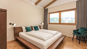 A bedroom with a wooden bed, two bedside lamps, a view of mountains through a window, and a green chair. - Panorama Dachstein by Interhome (Rohrmoos)