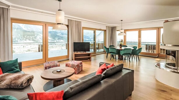 A spacious living area with a sofa, coffee table, and dining table. There is a television and a fireplace. Large windows offer a view of mountains. - Panorama Dachstein by Interhome (Rohrmoos)
