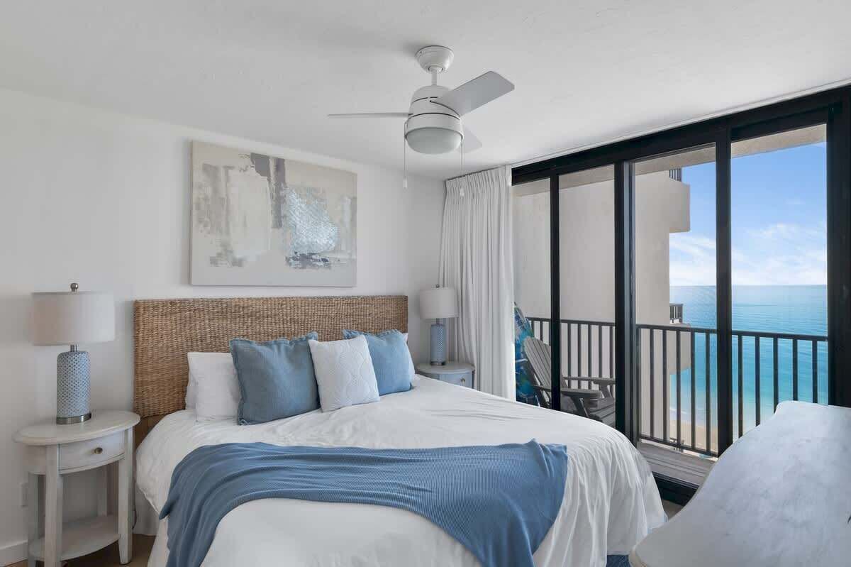 A hotel room with a bed, a ceiling fan, a large window with a view of the ocean, and a balcony with a chair.