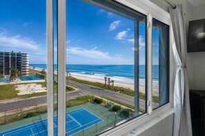 Vista desde una ventana sobre una playa, una carretera y una cancha de tenis.