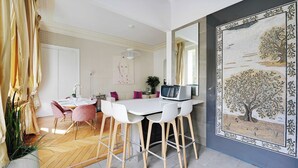 Een moderne keuken met witte kastjes, een eethoek met een witte tafel en roze stoelen, en een grote decoratieve muurschildering.