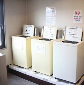Drei weiße Geräte, möglicherweise Waschmaschinen oder Trockner, mit Bedienungsanleitungen darauf, in einem Raum mit beigen Wänden und einem Fenster.