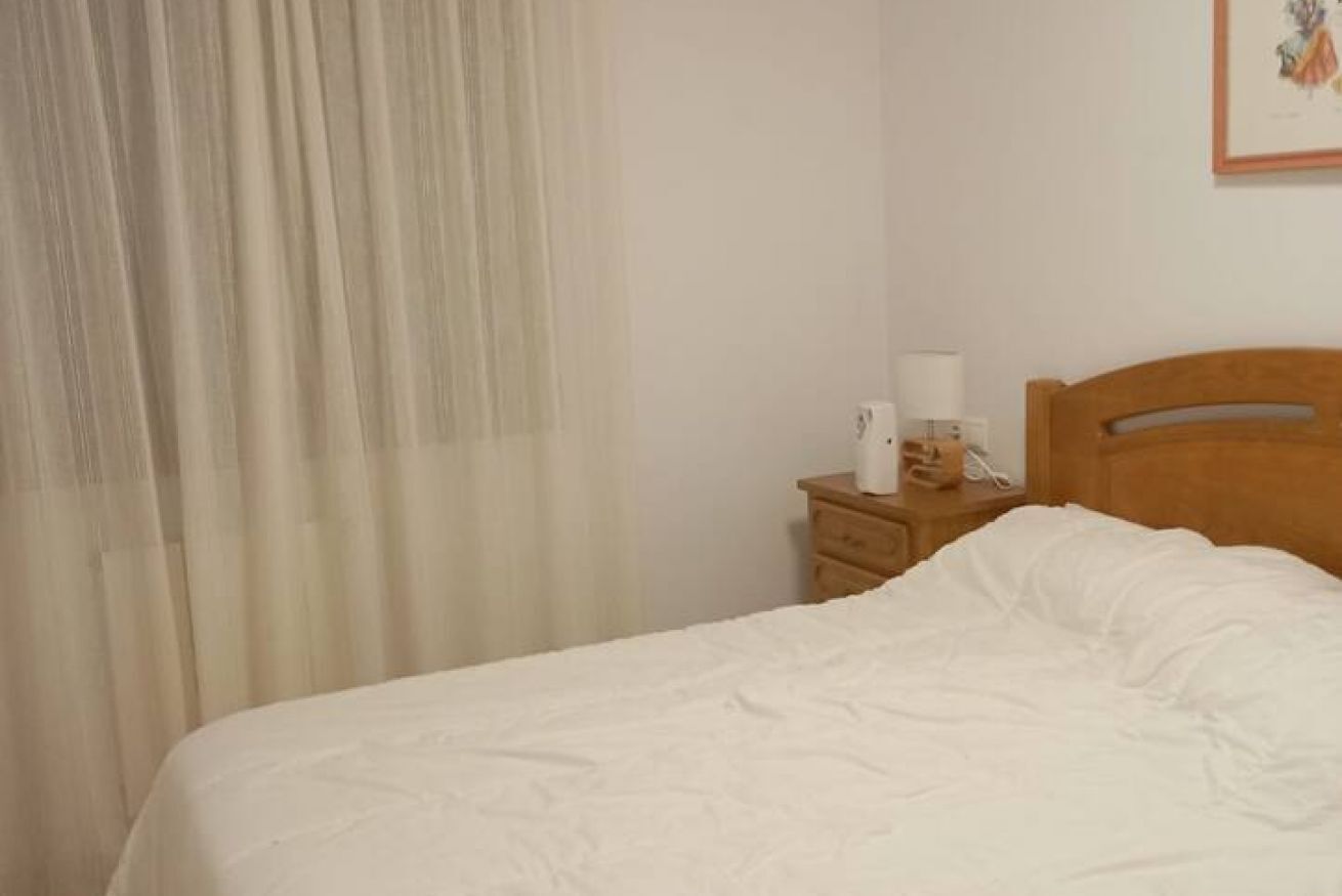 A neatly made bed with white bedding, a wooden nightstand with a lamp and a telephone, and a window with sheer curtains.