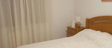 A neatly made bed with white bedding, a wooden nightstand with a lamp and a telephone, and a window with sheer curtains.