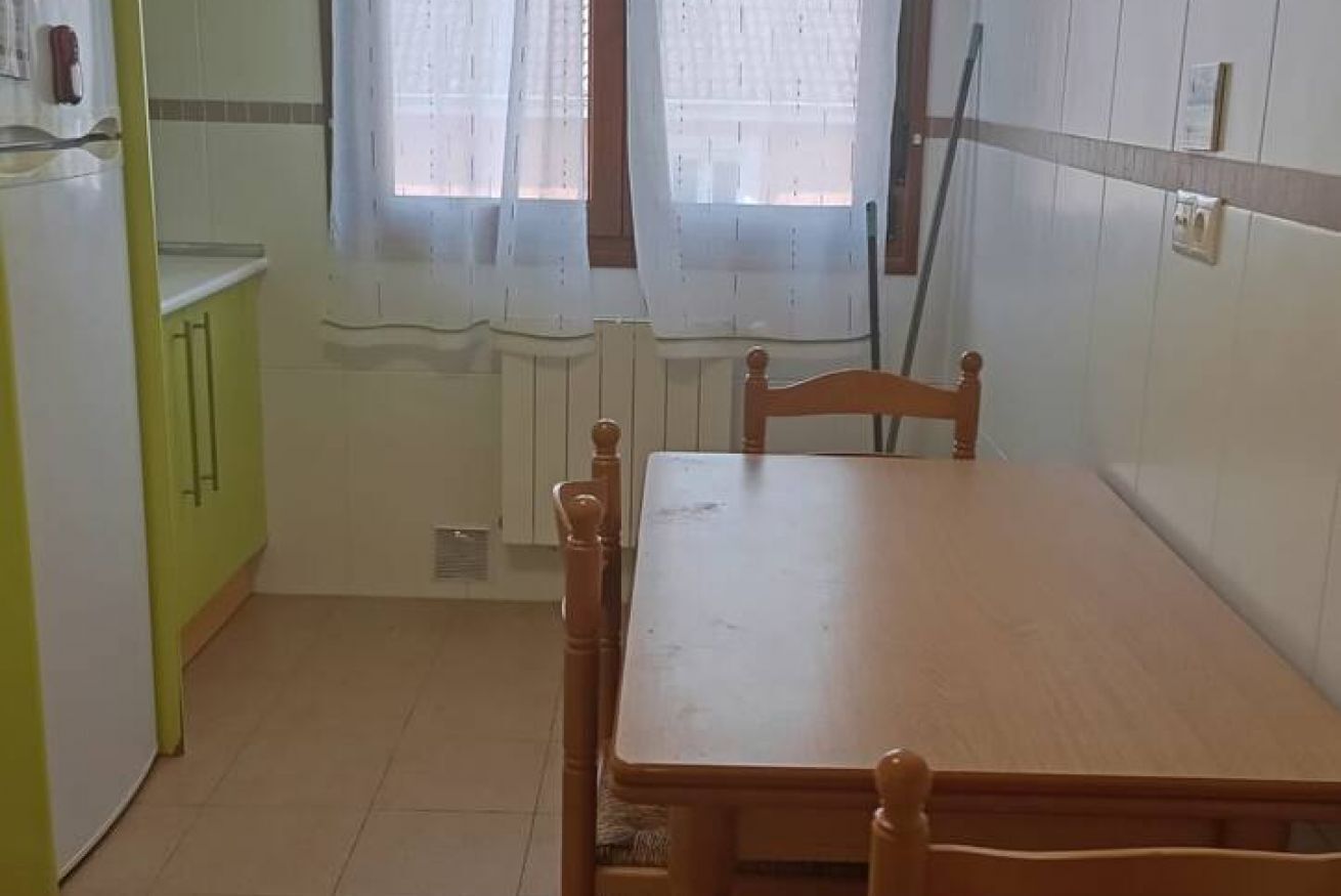 A kitchen with a wooden table and chairs, a refrigerator, and a window with curtains.
