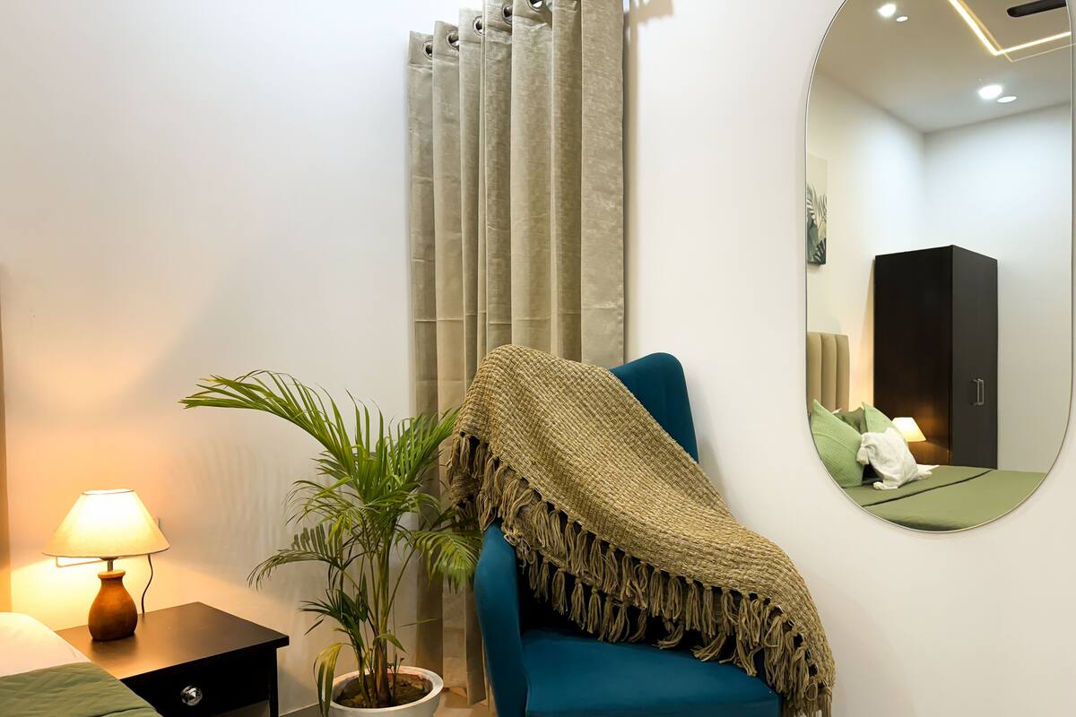 A modern bedroom with a teal sofa, a round mirror, a bedside table with a lamp, and a potted plant.