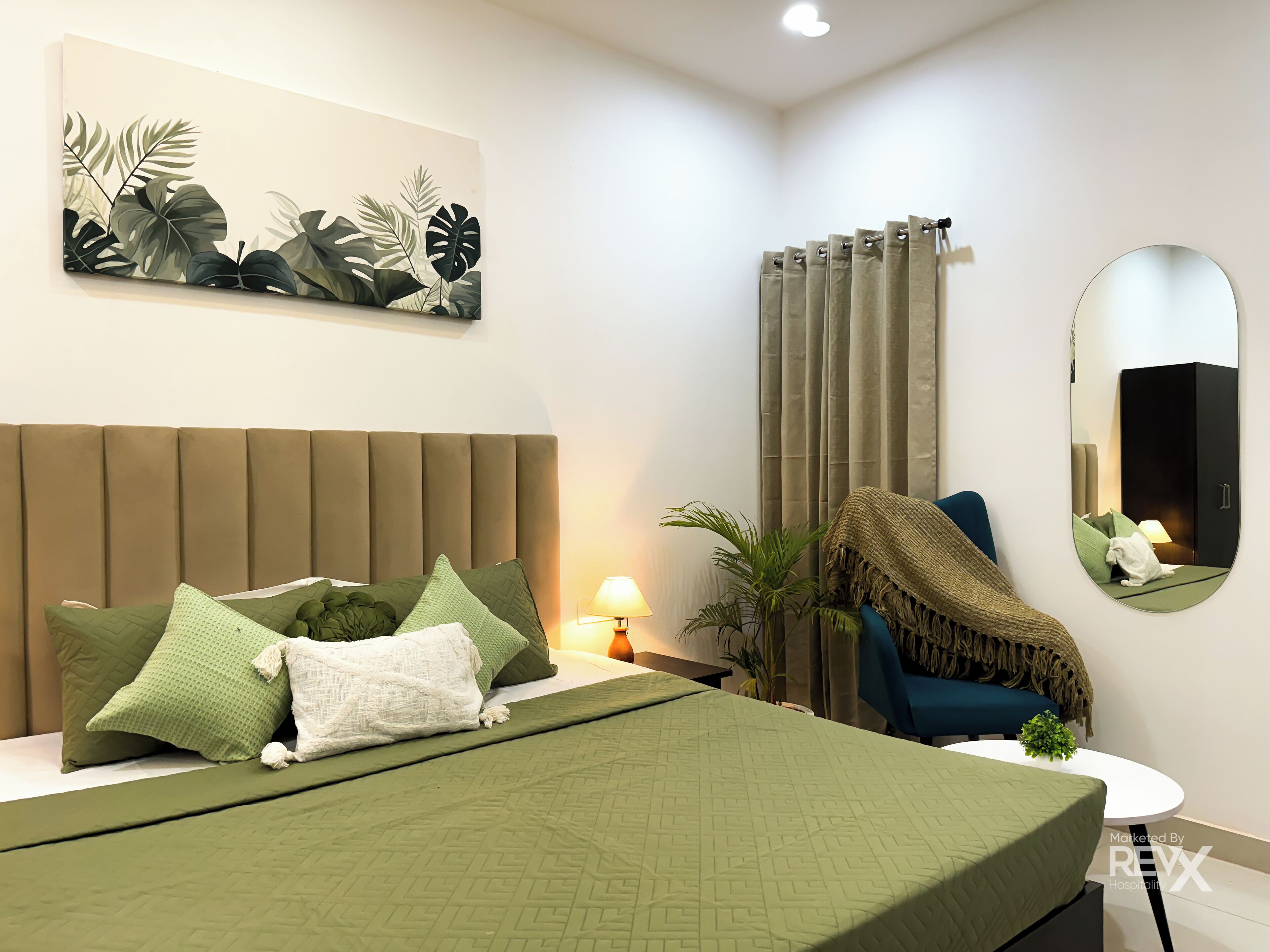 A modern bedroom with a large bed, green bedding, a headboard, a wall-mounted artwork, a bedside lamp, a mirror, a chair, and a small table with a plant.