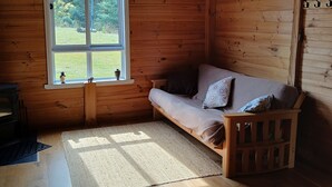 A cozy cabin interior with a wooden floor, a sofa with cushions, a window with a view of the outdoors, and a hanging light fixture. - cosy cottage with wood heater, surrounded by bushland (Middleton)
