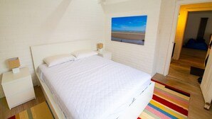 Ein Schlafzimmer mit einem Bett, einem Nachttisch, einer Lampe, einem farbenfrohen Streifen Teppich und einem Meeresblick-Gemälde an der Wand.