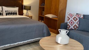 A hotel room with a large bed, a sofa, a wooden bookshelf, and a small table with a white pitcher.